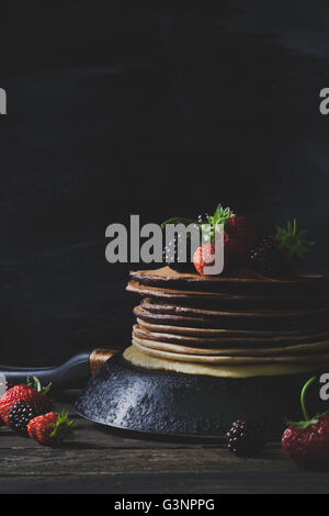 Chocolate pancakes in rusty pan with organic fruits like strawberries and blackberries on old wooden table Stock Photo