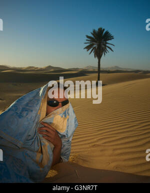 A local man sits on a sand dune with silky blue and gold fabric wrapped around his head and face to protect from blowing sand Stock Photo