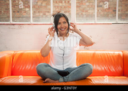 woman sitting crossed legged on a sofa listening to music on headphones on her ipad Stock Photo