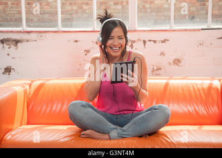 woman sitting crossed legged on a sofa listening to music on headphones on her ipad Stock Photo