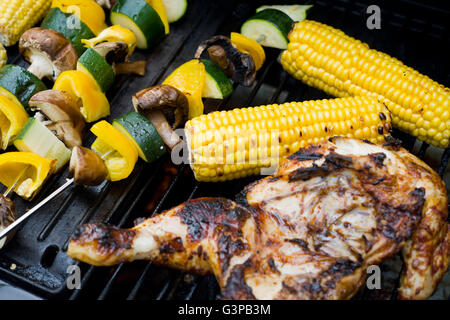 Spatchcock chicken on a barbecue with corn, vegetable skewers, burgers and coiled sausages. Stock Photo