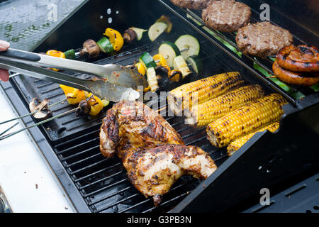 Spatchcock chicken on a barbecue with corn, vegetable skewers, burgers and coiled sausages. Stock Photo