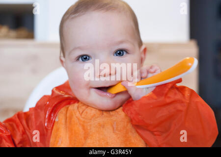 8 month baby boy having his cereal mush. He is wearing an orange bib. Trying to eat independently Stock Photo