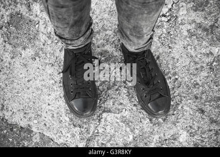 Teenager fashion theme, feet in black gumshoes and gray jeans standing on rough concrete floor