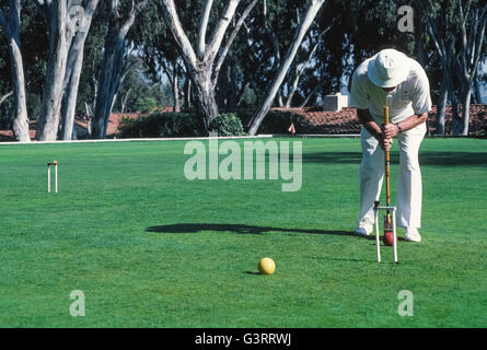 The traditional game of croquet is a popular activity for members of the the local croquet club who dress in all white apparel, as well as for guests at The Inn at Rancho Santa Fe, both of whom play on the sprawling front lawn of that luxury hotel in San Diego County, California, USA. Club members follow the official rules of the U.S. Croquet Association for Six-Wicket American Croquet that specify the equipment to be used, including the mallets, balls, wickets and stake. Here a male club player aims to strike his red ball with his mallet through a wicket. Stock Photo