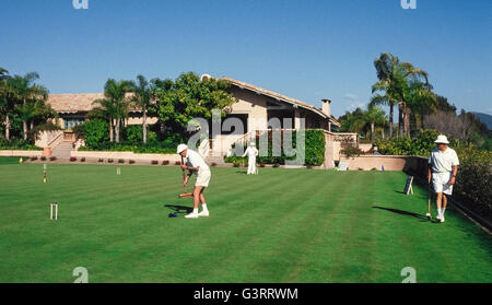 The traditional game of croquet is a popular activity for members of the the local croquet club who dress in all white apparel, as well as for guests at Rancho Valencia Resort & Spa, both of whom play on the sprawling croquet lawn of that luxury hotel in San Diego County, California, USA. Club members follow the official rules of the U.S. Croquet Association for Six-Wicket American Croquet that specify the equipment to be used, including the mallets, balls, wickets and stake. Here a male club player gets set to strike his blue ball with a square wooden mallet to drive an opponent's black ball. Stock Photo