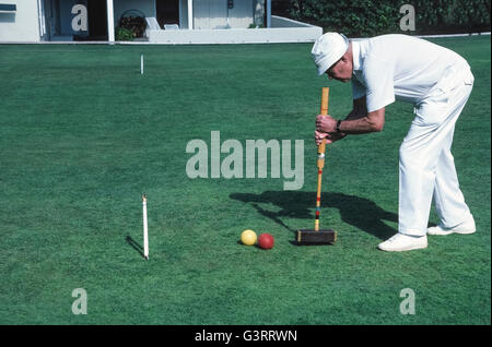 The traditional game of croquet is a popular activity for members of the the local croquet club who dress in all white apparel, as well as for guests at The Inn at Rancho Santa Fe, both of whom play on the sprawling front lawn of that luxury hotel in San Diego County, California, USA. Club members follow the official rules of the U.S. Croquet Association for Six-Wicket American Croquet that specify the equipment to be used, including the mallets, balls, wickets and stake. Here a male club player strikes his red ball with a square wooden mallet to drive away an opponent's yellow ball. Stock Photo