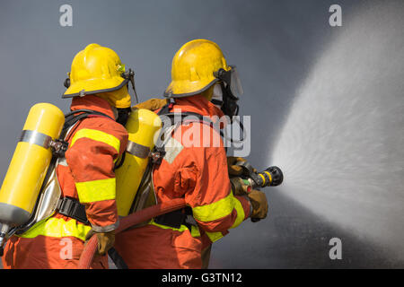 2 firefighters spraying water in fire fighting with dark smoke background Stock Photo