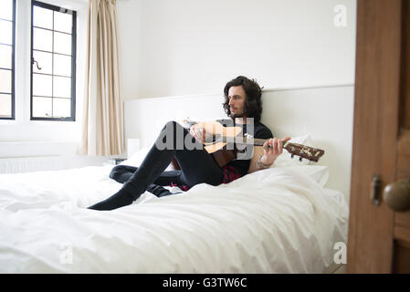 A cool young man laying on a bed with a guitar. Stock Photo