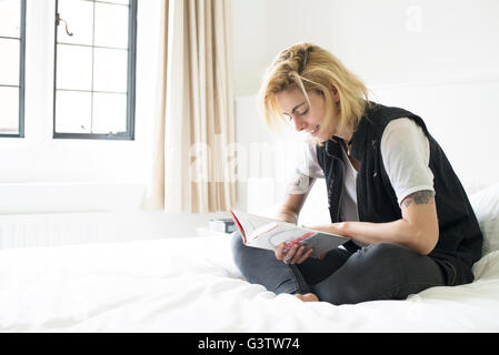 A young woman with tattooes sitting on a bed reading. Stock Photo