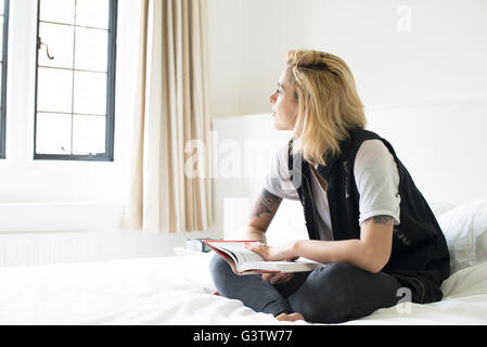 A young woman with tattooes sitting on a bed reading. Stock Photo