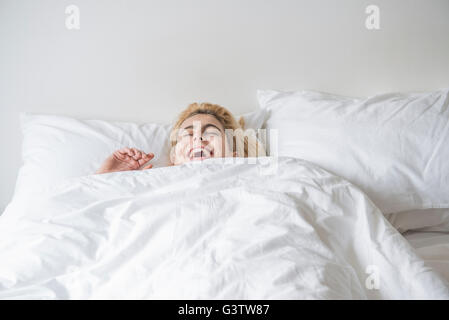 A cool young woman lying in a bed with he covers pulled up to her chin and laughing. Stock Photo