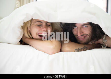 A cool young tattooed couple peeking out from under a duvet on a bed. Stock Photo