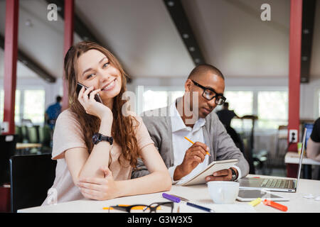 Cheerful beautiful young business woman talking on cell phone and working with serious african man in office Stock Photo