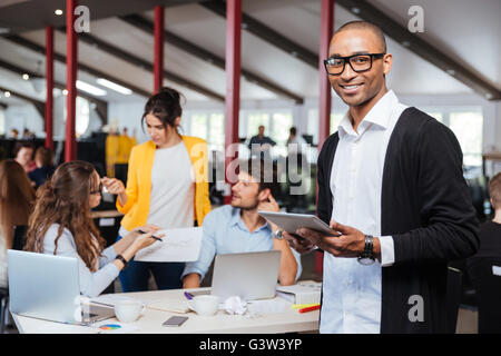 Cheerful african young businessman using tablet in office with colleagues on background Stock Photo