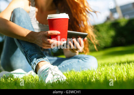 Young woman with red disposable paper cup of coffee using tablet pc on a green lawn in a park Stock Photo