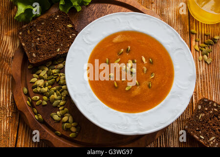 Pumpkin and carrot soup with seeds in a white plate on the wooden background. Green lettuce and olive oil. Stock Photo