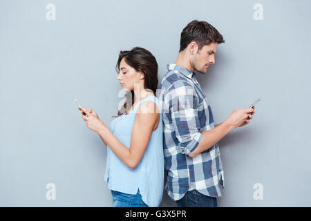 Portrait of young couple standing back to each other and looking at the phone isolated on gray background Stock Photo