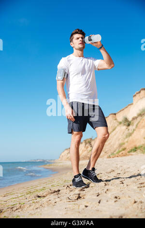 Handsome young sportsman standing and drinking water on the beach Stock Photo