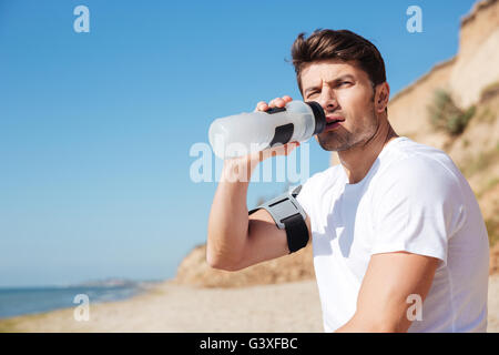 Serious young sportsman sitting and drinking water on the beach Stock Photo
