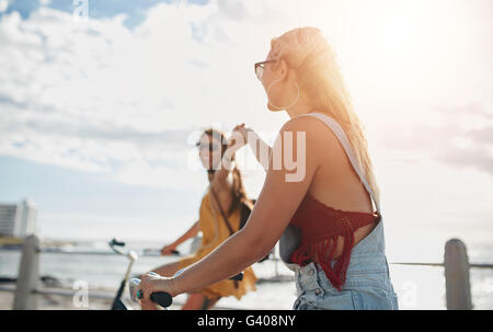 Female friends riding cycles on the seaside promenade and having fun. Young women enjoying riding bicycles on a summer day. Stock Photo