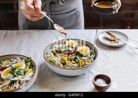 A woman is drizzling a veggie bowl with warm peanut dressing photographed from the front view. Stock Photo