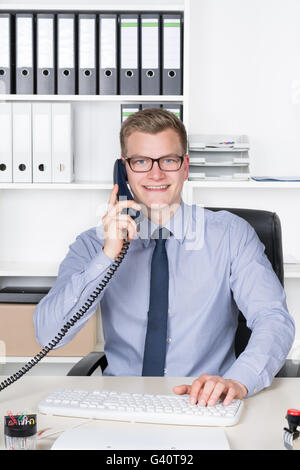 Young smiling businessman with glasses is phoning and typing at the computer keyboard while sitting at the desk in the office. A Stock Photo