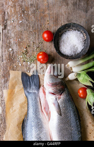 Two raw uncooked seebass fish on baking paper with sea salt, dry herbs and vegetables over old wooden background. Top view Stock Photo