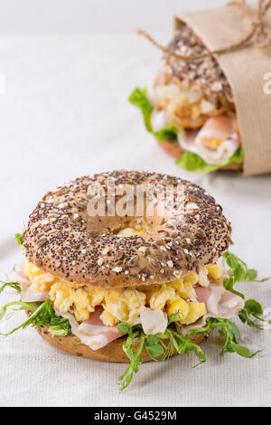 Whole Grain bagels with scrambled eggs, pea sprout and prosciutto ham on wood plate over wooden textured background. Stock Photo