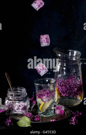 Ice cubes with flowers falling into glass of lilac lemonade with lemon. Glass jar of sugared lilac flowers and glass pitcher on Stock Photo