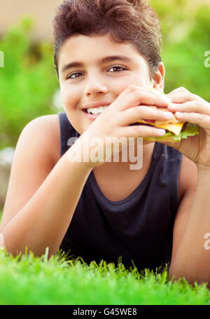 Portrait of a cute happy teen boy lying down on a fresh green grass field and eating tasty burger, enjoying fastfood Stock Photo