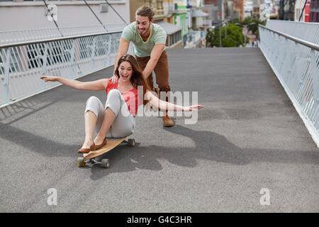MODEL RELEASED. Young woman sitting on skateboard with man pushing. Stock Photo