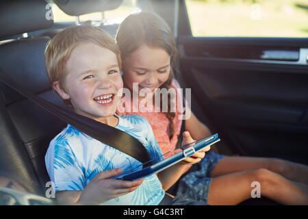 MODEL RELEASED. Brother and sister in the back seat of the car wearing seat belts, using a digital tablet. Stock Photo