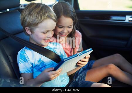 MODEL RELEASED. Brother and sister in the back seat of the car wearing seat belts, using a digital tablet. Stock Photo