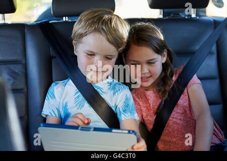 MODEL RELEASED. Brother and sister in the back seat of the car wearing seat belts, using a digital tablet. Stock Photo