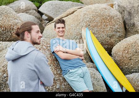 MODEL RELEASED. Young man leaning on rocks with surfboard. Stock Photo