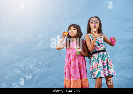 Portrait of adorable little girls blowing soap bubbles against blue wall. Two young girls playing together. Stock Photo