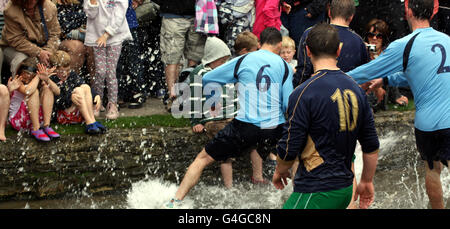 Two young children take cover from the water as Bourton Rovers 1st XI (light blue) battle for possession with Bourton Rovers 2nd XI (dark blue) during the annual Football in the River match in Bourton-on-the-Water, Gloucestershire on Bank Holiday Monday. The teams of local players battle it out in the annual event which takes place in the River Windrush in centre of town. Stock Photo