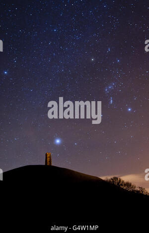 Photo. Looking south over the Somerset sky, low and fast moving clouds reflect light pollution from street lighting, as the brightest star in the night sky, Sirius (the Dog Star) rises low above St. Michael's Tower on Glastonbury Tor with the constellation of Orion (middle right) clearly visible. Stock Photo