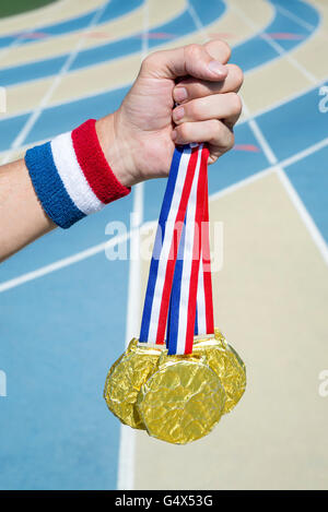 Hand of American athlete holding gold medals hanging from USA colors red, white, and blue ribbon against a running track Stock Photo