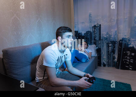 Fasionable young couple spend their free time at home, where young man  playing a video game holding a console while his girlfriend lying on the sofa and surf the Internet by her mobile phone. Stock Photo