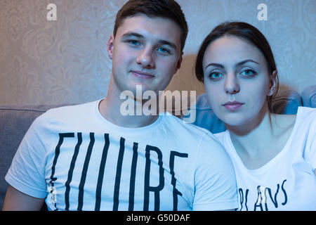 Portrait of young and attractive couple sitting on a couch together in front of a TV at home in a relaxed atmosphere and watching television. Stock Photo