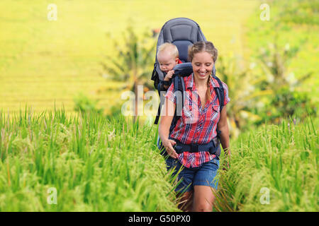 Nature walk in green rice terrace field. Happy mother hold little traveller in carrying backpack. Baby ride on woman back.
