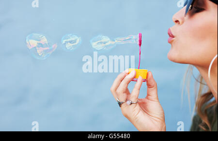 Close up side view shot of young female model blowing soap bubbles on blue background. Focus on hands and wand. Stock Photo