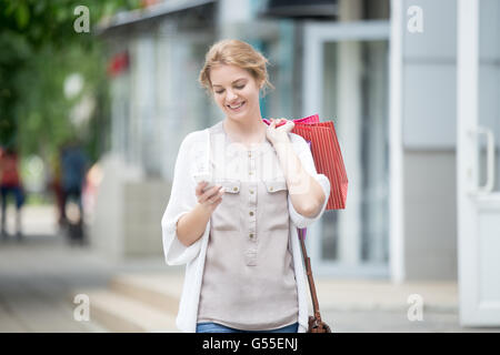 Happy beautiful person looking at phone while carrying shopping bags with purchases. Young joyful model reading message, texting Stock Photo