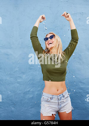 Portrait of beautiful young woman throwing glitters on herself while standing against blue wall Stock Photo