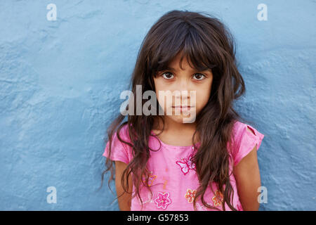 Close up portrait of innocent little girl standing against blue wall, she is looking at camera with serious expression on her fa