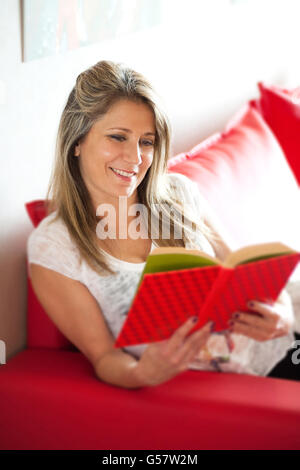 Single smiling gorgeous mature woman in white blouse enjoying a book reading session while seated on red sofa indoors Stock Photo