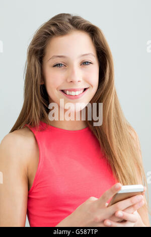 Portrait of a teenage girl using a smartphone. She is smiling at the camera against a plain background. Stock Photo