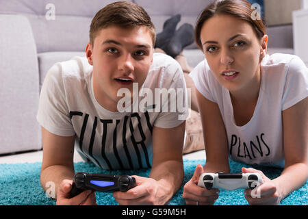 Young attractive couple lying on a rug in their living room playing video games looking towards the camera and pointing with fascinated expressions. Stock Photo